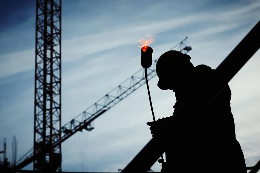 Silhouette of a worker holding a lit gas torch against a sky with cranes behind