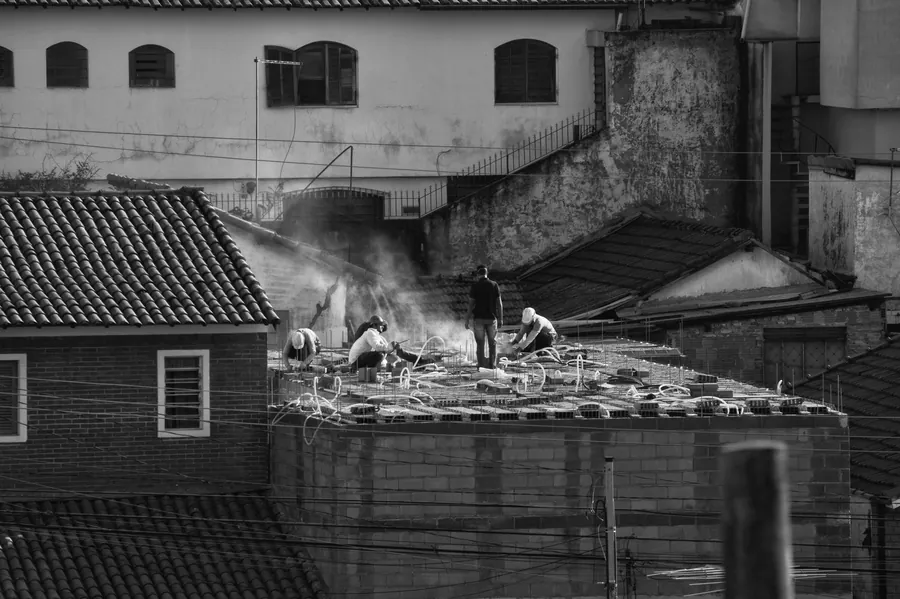 Black and white photo of workers laying a new covering on a flat roof among older tiled roofs