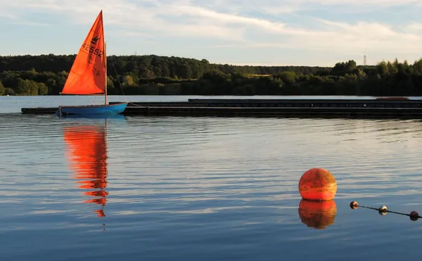 Red-sailed dinghy on a still lake with a buoy reflected in the water